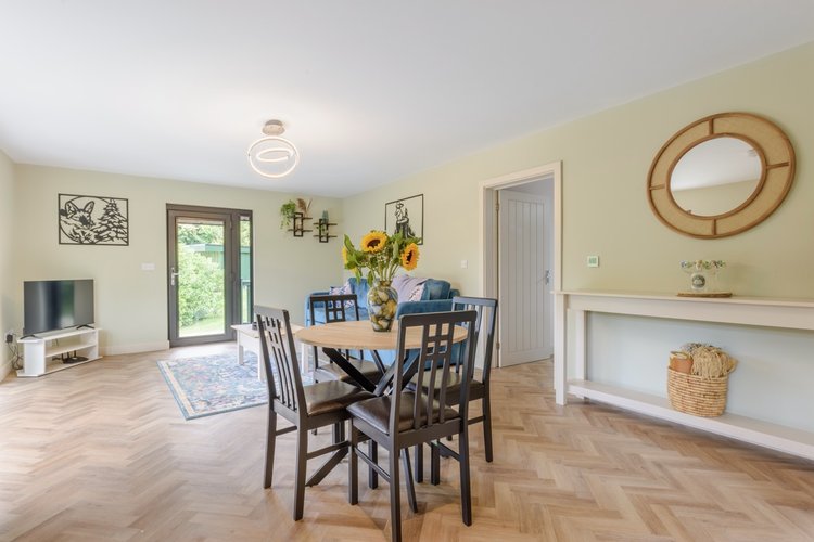 The dining area, with its round table and chairs, features a vibrant sunflower centerpiece. A TV sits nearby, while the large window offers a sweeping view of Rowley Meadow's lush greenery.