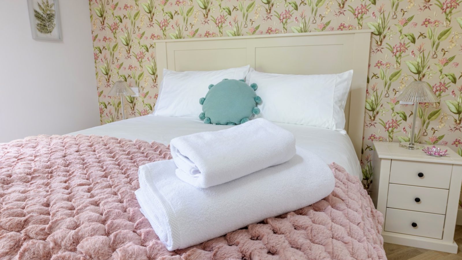 A neatly made bed with a pink textured blanket awaits at Rowley Meadow, floral wallpaper, and towels completing the look.