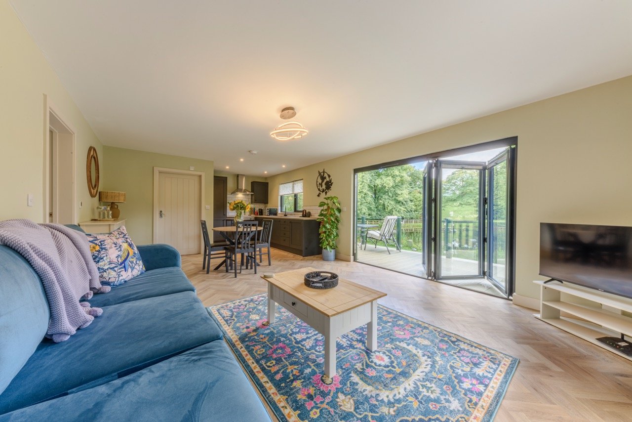 The spacious living room at Rowley Meadow features a blue sofa, patterned rug, wooden coffee table, and TV. Open doors lead to a patio with lush greenery, creating the perfect blend of indoor comfort and outdoor charm.