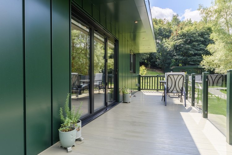 The modern outdoor patio, featuring Rowley Meadow's signature green paneling and glass railing, is adorned with potted plants. It offers a serene view of the garden below, complete with trees and a small lawn.