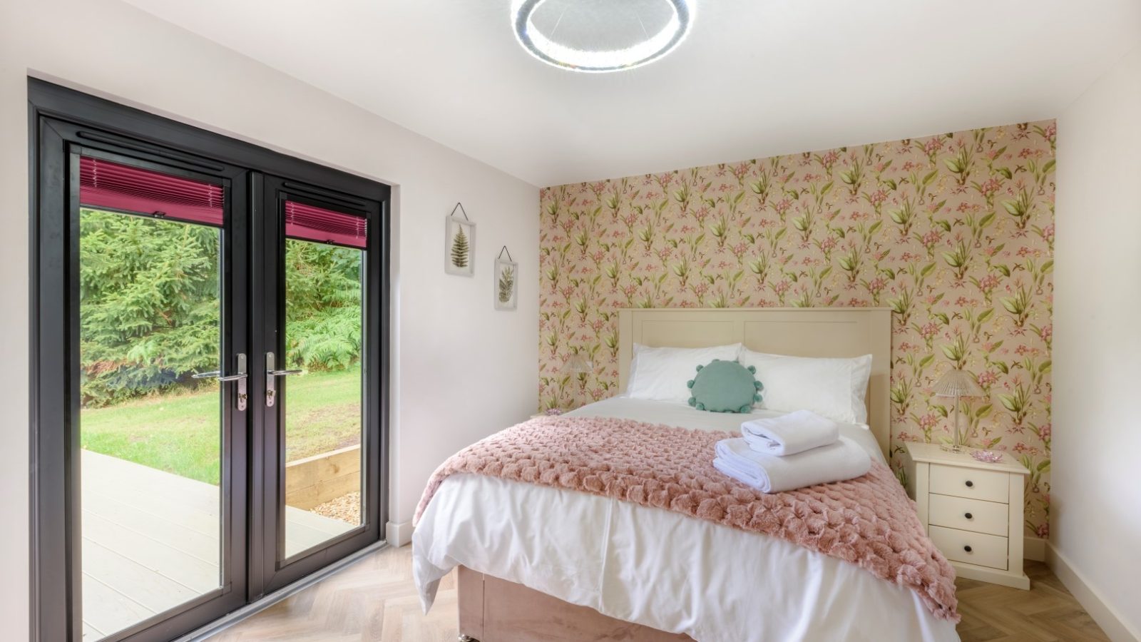 Bedroom with floral wallpaper, double bed, and pink throw. Towels folded neatly. Glass doors open to a Rowley Meadow view.