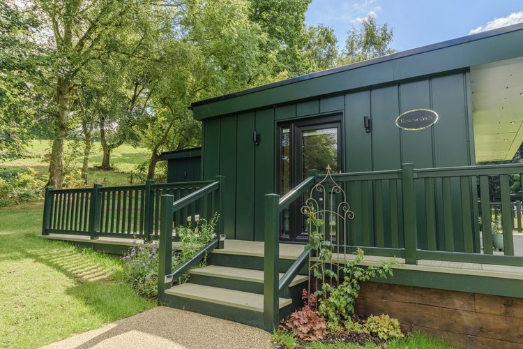 Nestled in the serene beauty of Rowley Meadow, this green cabin with stairs and a railing is surrounded by trees and grass under a clear blue sky.