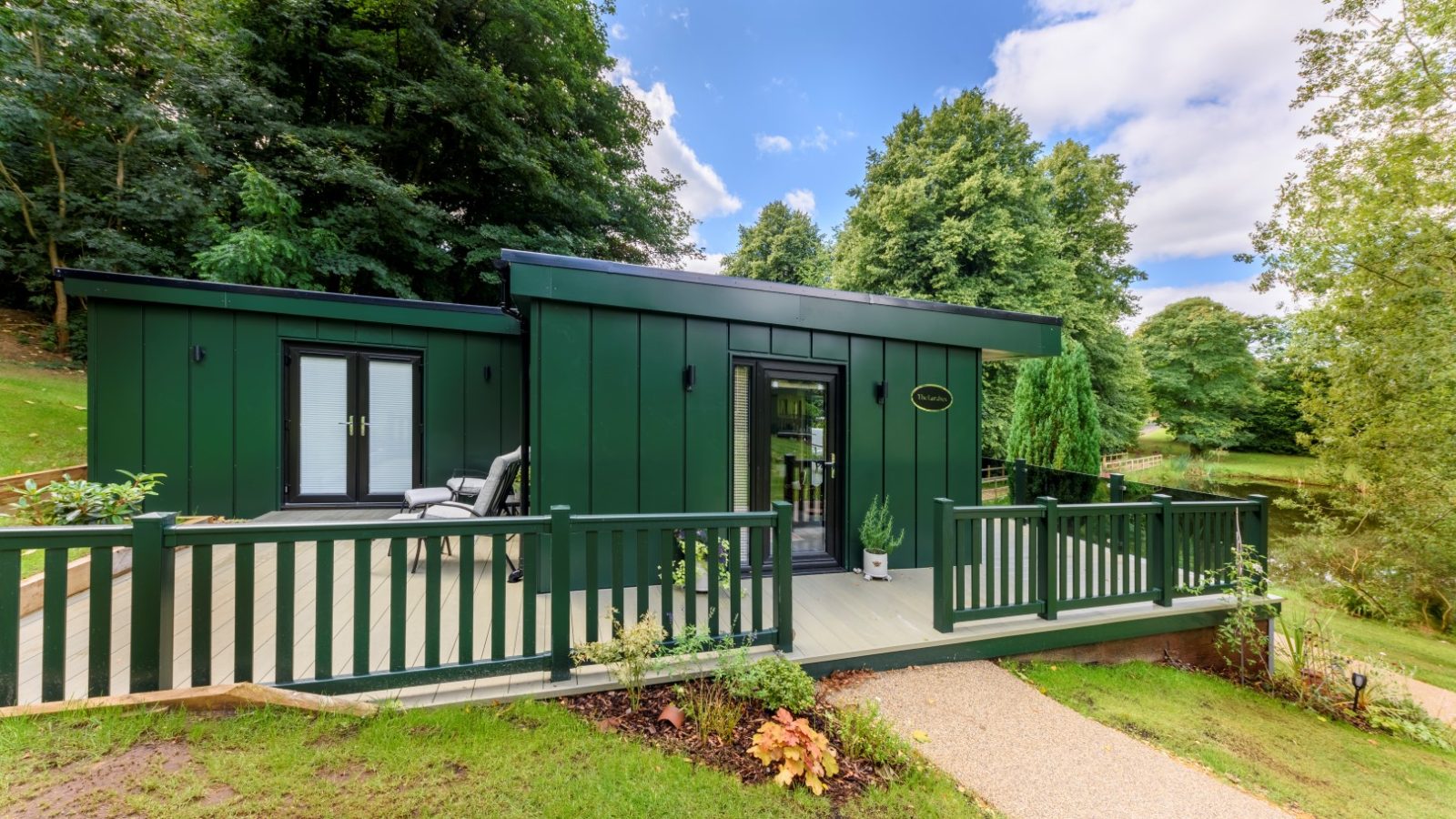 Modern green cabin with wooden deck in Rowley Meadow, surrounded by trees and grass under a blue sky with clouds.