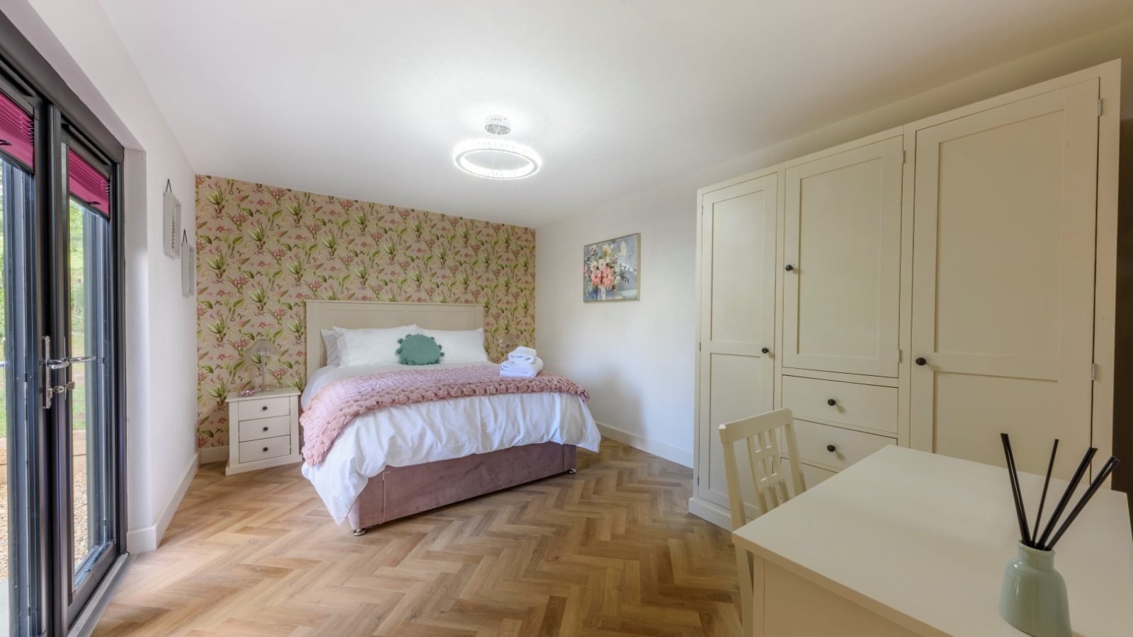 Bedroom with a bed, two bedside tables, and a wardrobe. Floral wallpaper and wood flooring echo Rowley Meadow charm.