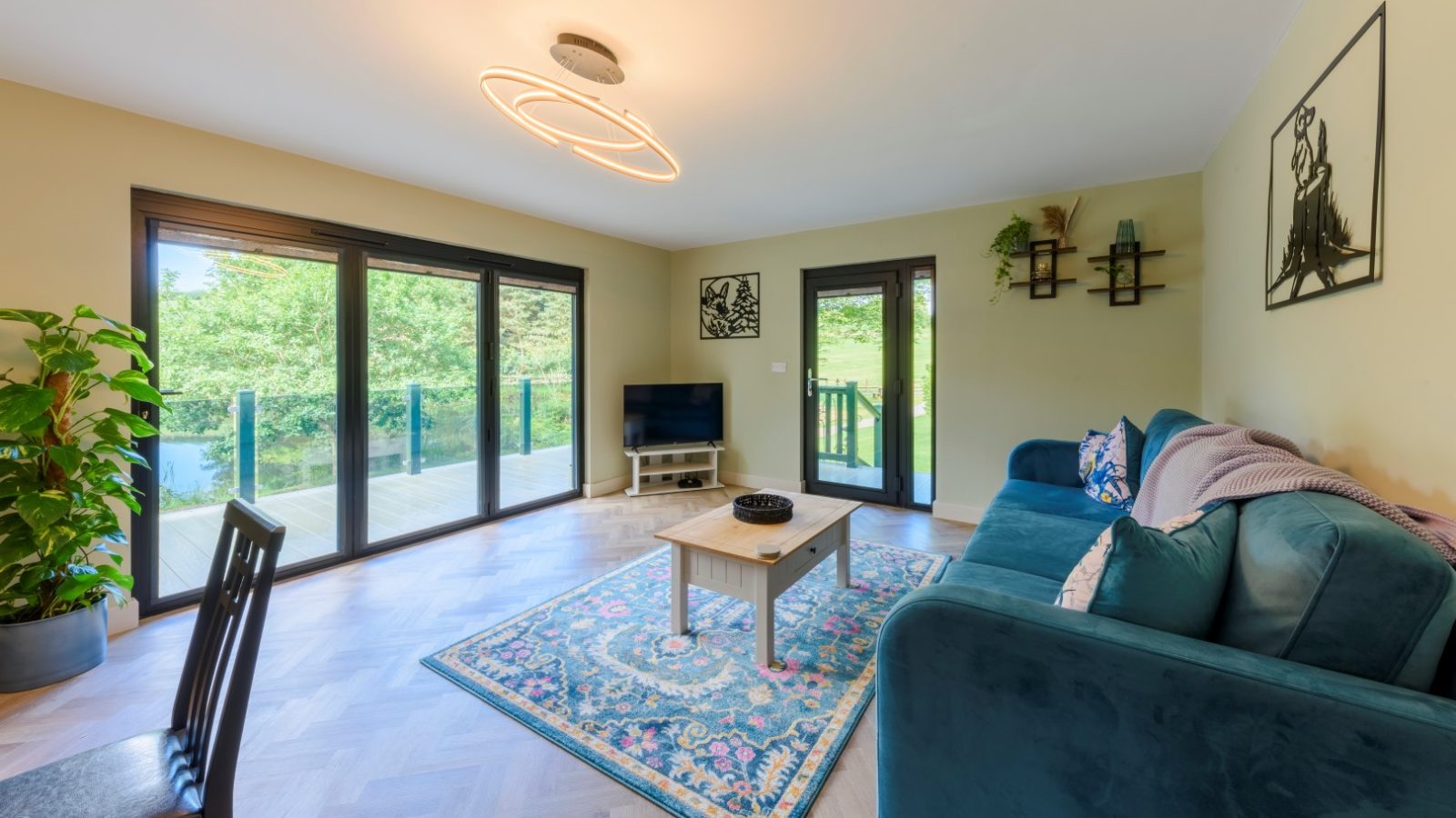 Modern living room with a large blue sofa, coffee table, and TV overlooking the deck at Rowley Meadow Lodges.