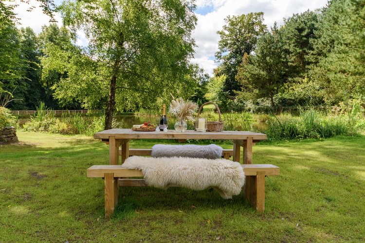 A picnic table with a sheepskin throw on a bench, set with food items, sits in Rowley Meadow. This idyllic spot by the pond is surrounded by lush trees and offers the perfect setting for a serene escape.