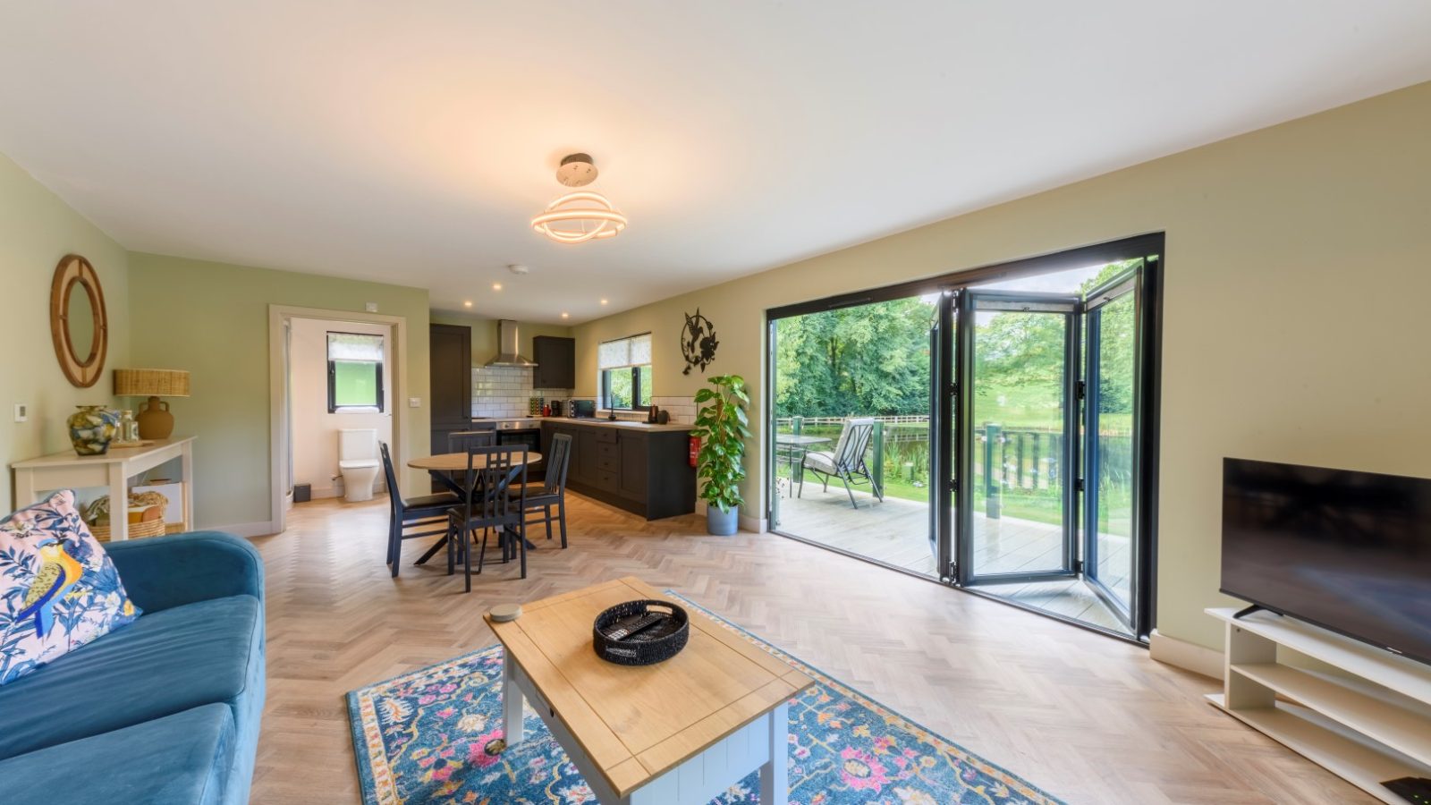 Open-plan living room with blue sofa and TV. Glass doors lead to a Rowley Meadow patio, offering a serene garden view.