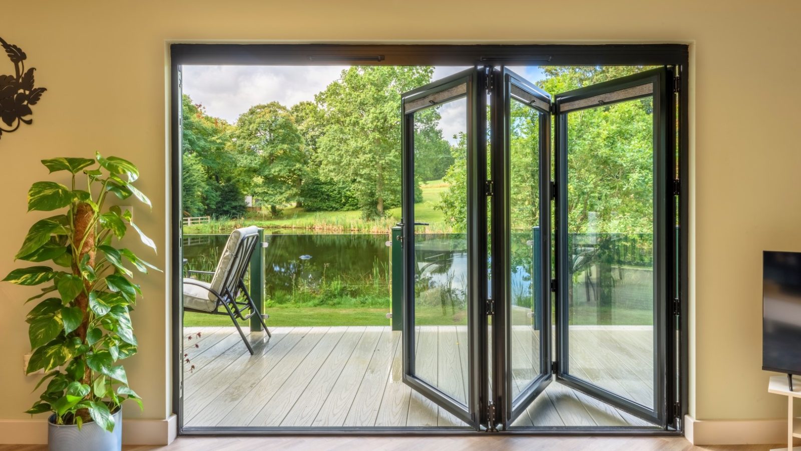 Open glass bifold doors to a deck with a chair overlooking Rowley Meadow's lake and trees. Indoor plant visible on the left.