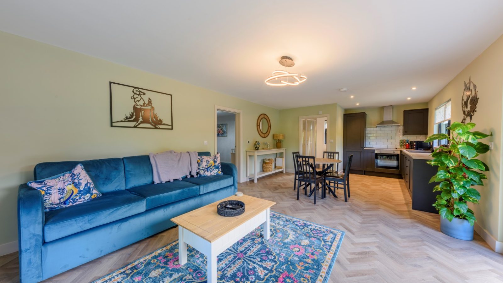 Living room with blue sofa, patterned rug, wooden coffee table, dining set, and an open kitchen at Rowley Meadow Lodges. Bright decor and plants.