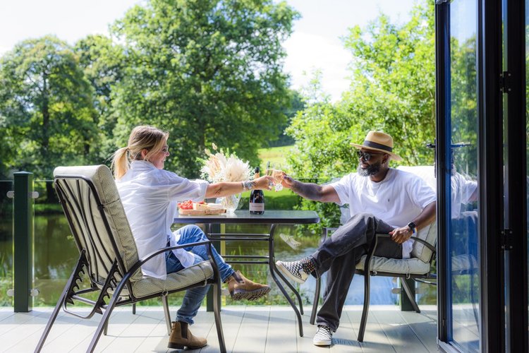 Two people toast with drinks at an outdoor table on a balcony overlooking the lush greenery of Rowley Meadow and shimmering water.