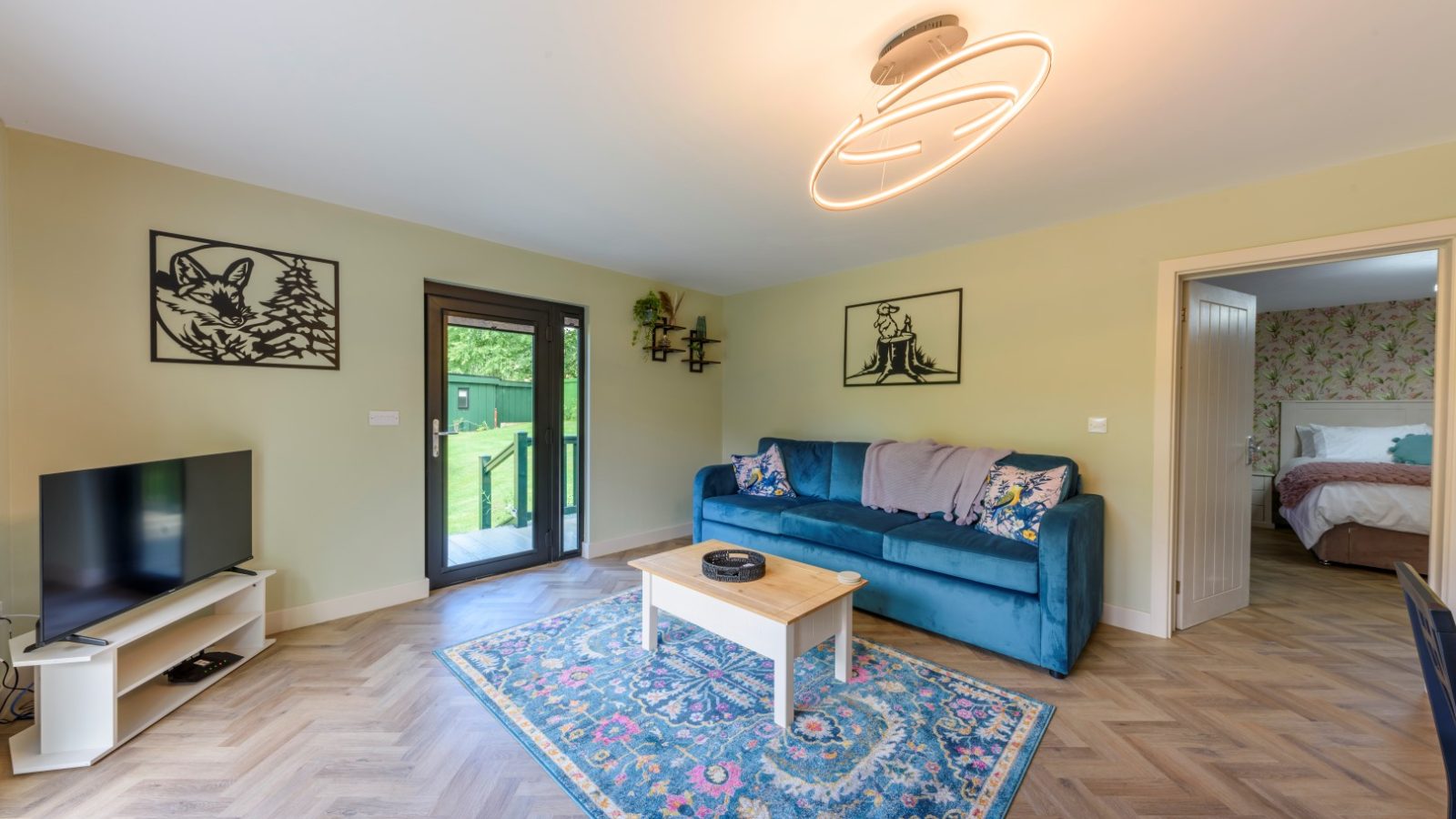 A living room with a blue sofa, wooden coffee table, flat-screen TV, and art on walls. A Rowley Meadow doorway leads to a bedroom.