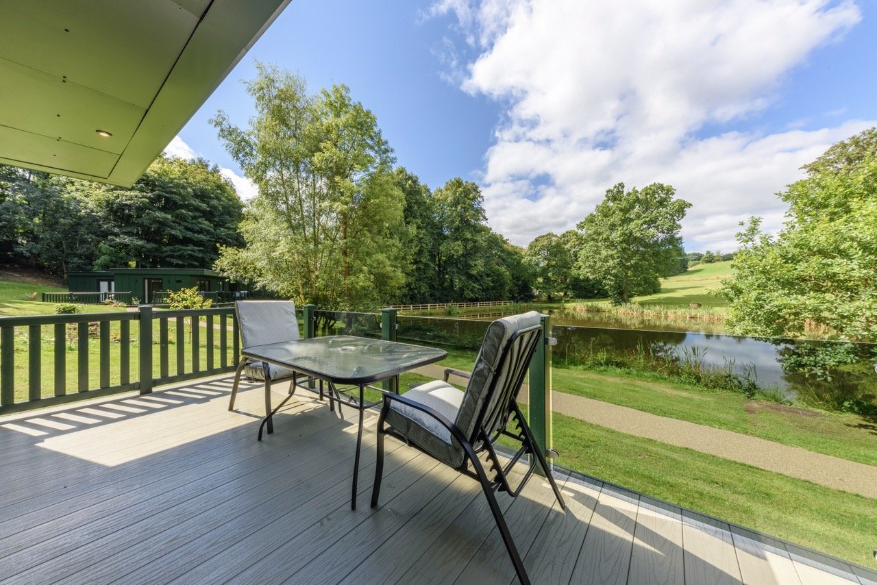 A patio with a glass table and chairs offers a serene view of Rowley Meadow, where lush greenery meets a tranquil pond beneath a partly cloudy sky.