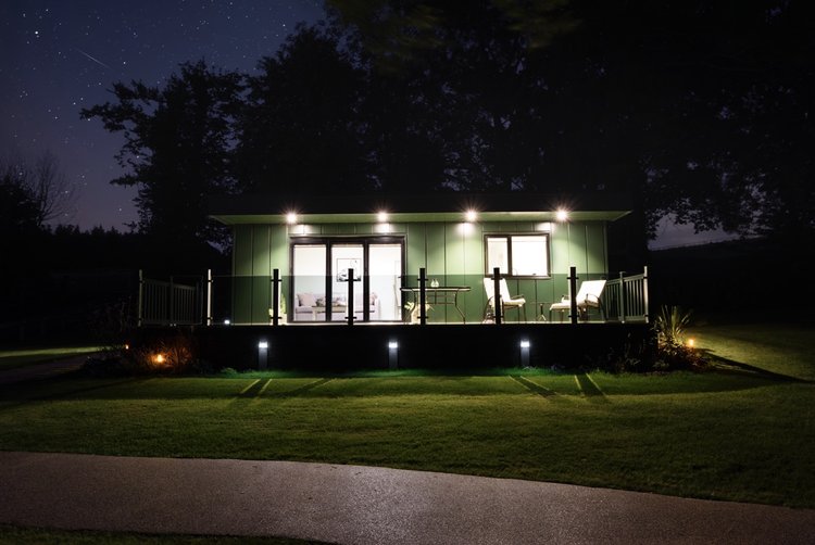 Small modern cabin with lit porch and large windows, nestled in Rowley Meadow, surrounded by grass and trees under a starry night sky.