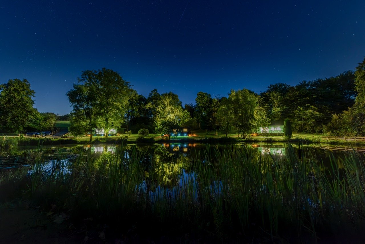 A serene night view of a calm pond surrounded by trees, with lights illuminating the scene under a clear starry sky at Rowley Meadow.