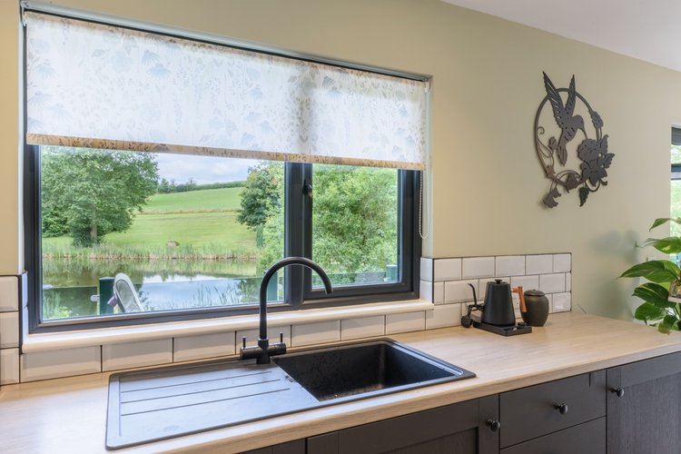 A kitchen sink with a black faucet overlooks the serene Rowley Meadow through a window adorned with white curtains.