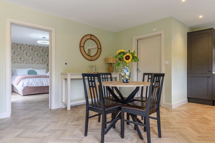 The dining area, nestled within Rowley Meadow, features a round table and four chairs. A vase of sunflowers adds charm, while the view reveals a bedroom adorned with floral wallpaper.