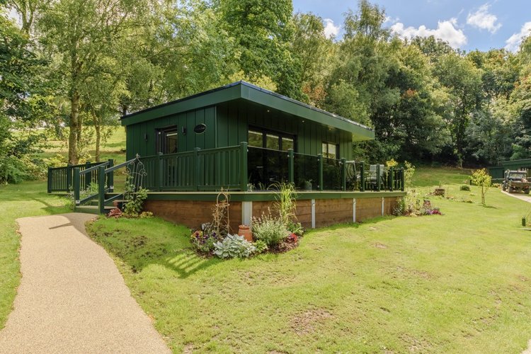 The Green Cabin at Rowley Meadow boasts large windows and a wraparound deck, nestled amidst lush grass and trees under the expansive blue sky.