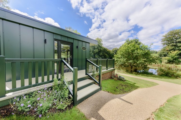 Nestled in Rowley Meadow, this green modular cabin features stairs surrounded by trees, with a path meandering to a small pond beneath a partly cloudy sky.