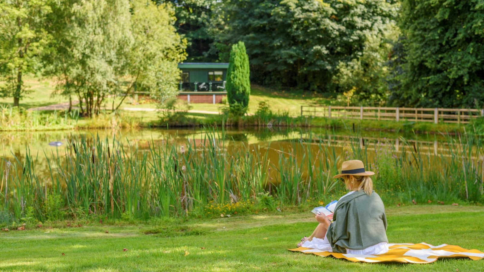 Person in a hat sits on a yellow blanket by Secret Waterside Lodges, reading a book, surrounded by trees and greenery.