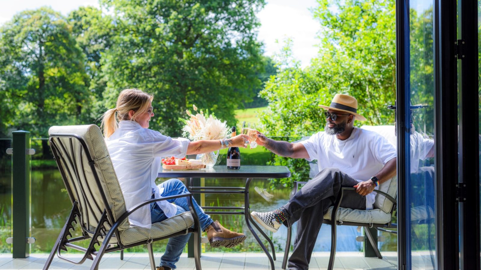 Two people sit on a patio by Rowley Meadow, toasting with glasses. A table with food and a bottle is between them on a sunny day.