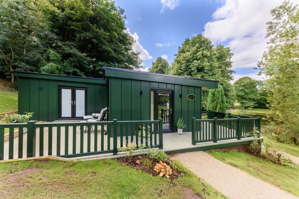 Nestled in the serene embrace of Rowley Meadow, this green modern cabin boasts a large deck, surrounded by trees and a clear sky in the background.