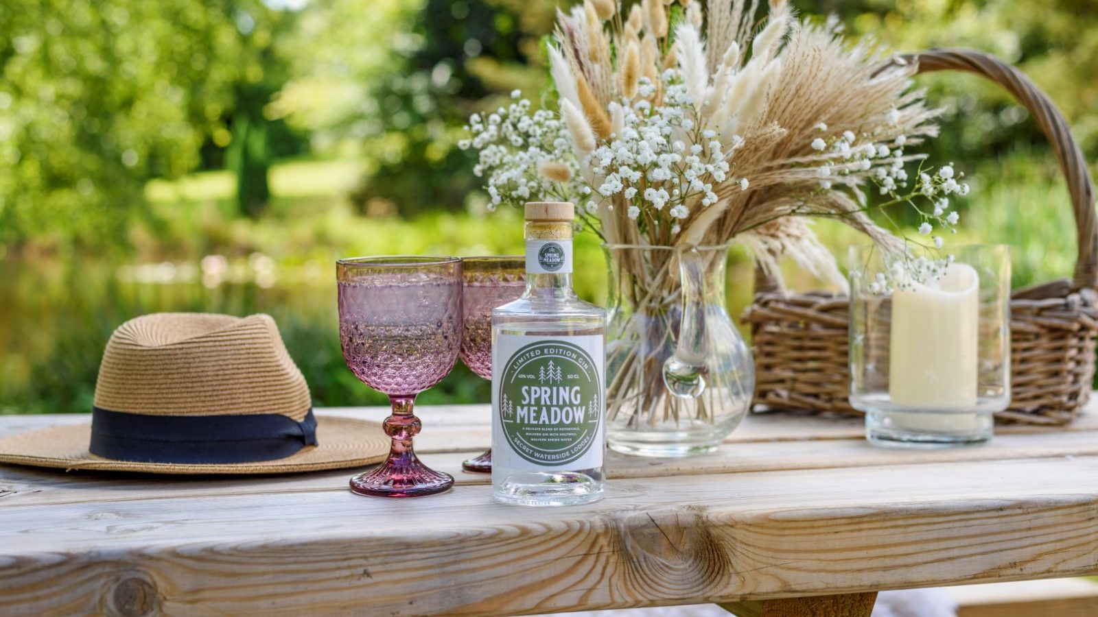 A rustic outdoor table with a hat, two glasses, Spring Meadow bottle, dried flowers, and candle by Rowley Meadow.