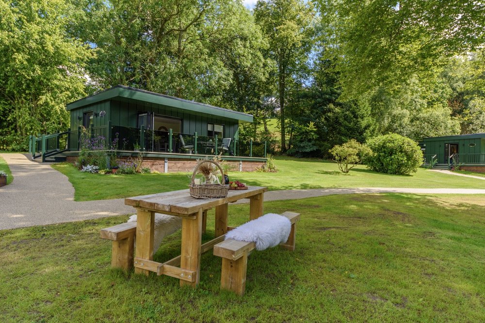 Nestled in the enchanting Rowley Meadow, this lodge is surrounded by a lush green setting. A wooden picnic table graces the foreground, with a charming basket perched on the bench, inviting you to unwind and savor the serene beauty.