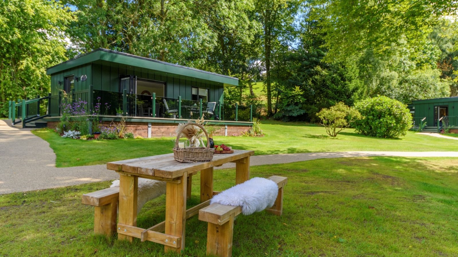 Wooden picnic table with a cushion and basket on Rowley Meadow, near a modern cabin surrounded by trees and greenery.