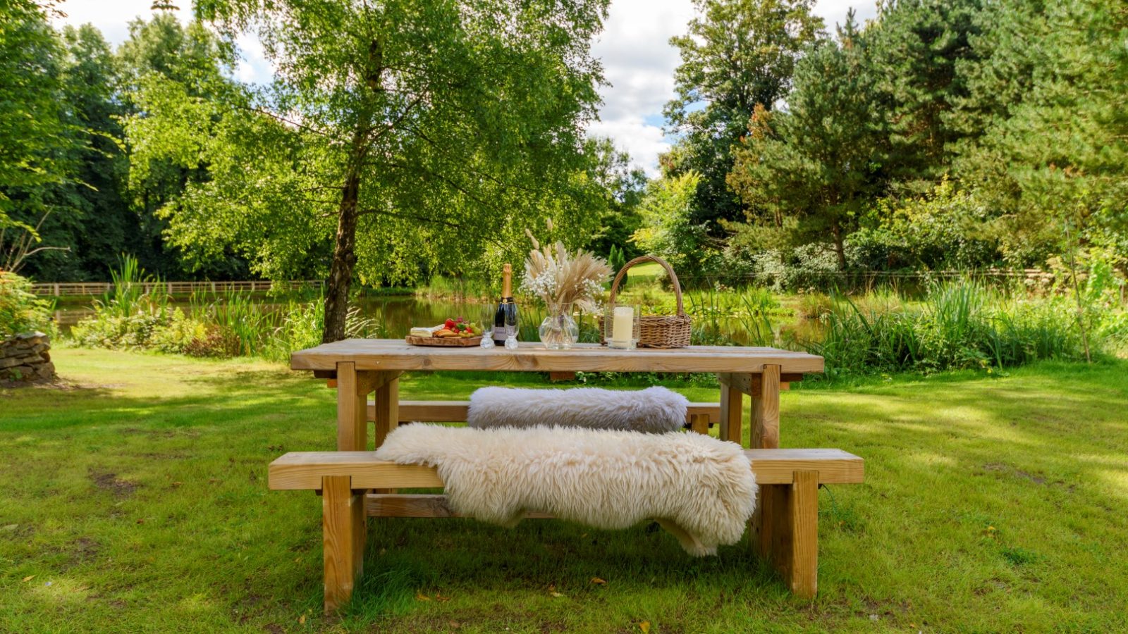 Wooden picnic table with plush blanket, wine, and cheese near Rowley Meadow's serene pond by Secret Waterside Lodges.