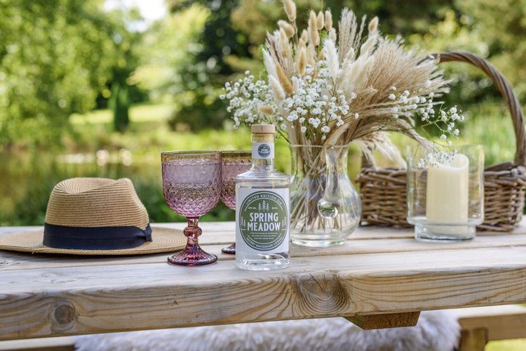 A straw hat, two glasses, and a Rowley Meadow bottle sit alongside a vase with flowers on a wooden table in an inviting outdoor setting.