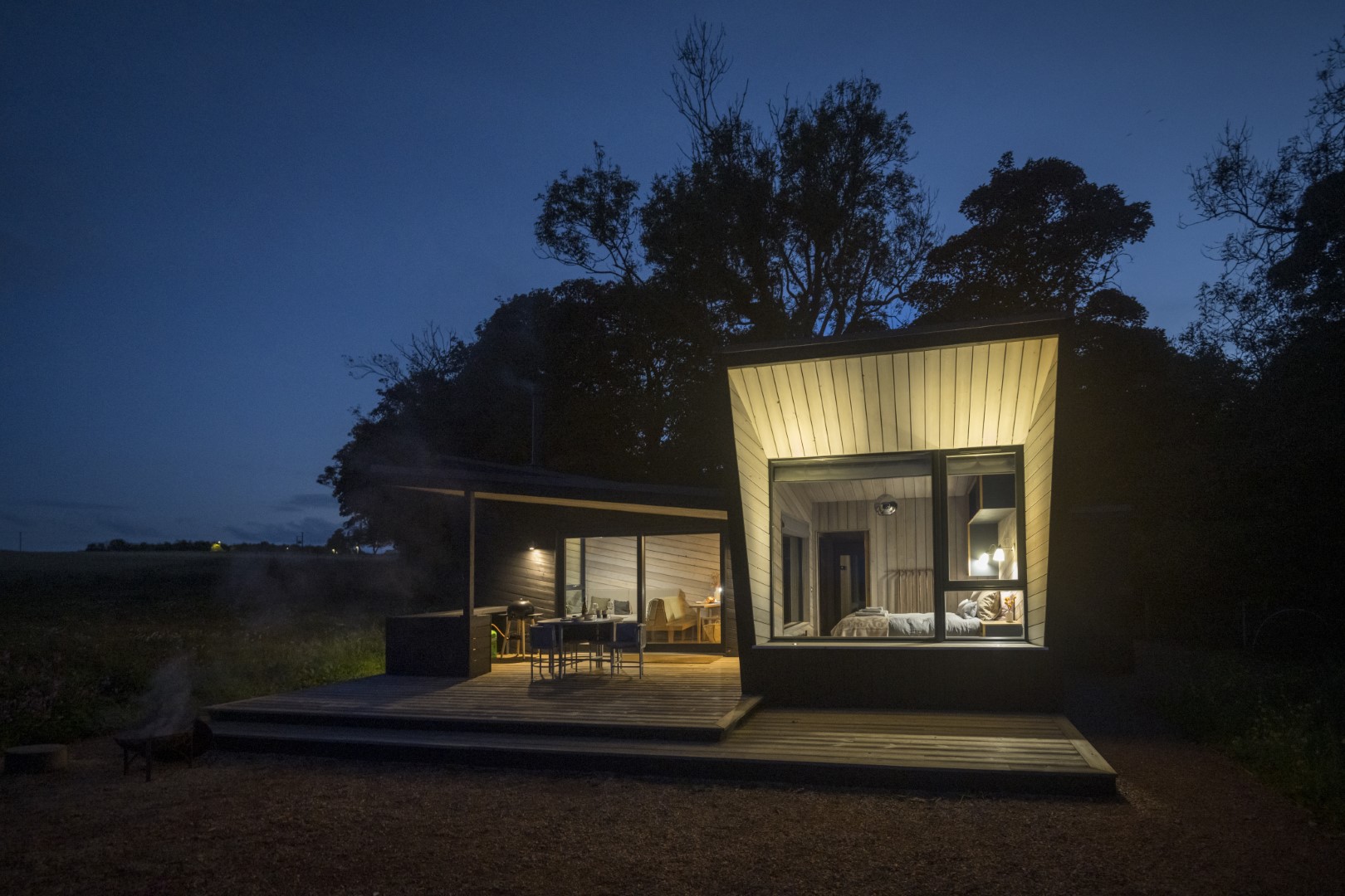 Modern cabin with large windows illuminated at night, surrounded by trees, featuring a spacious deck with outdoor seating ideal for enjoying summer holiday activities and breaks. Places to