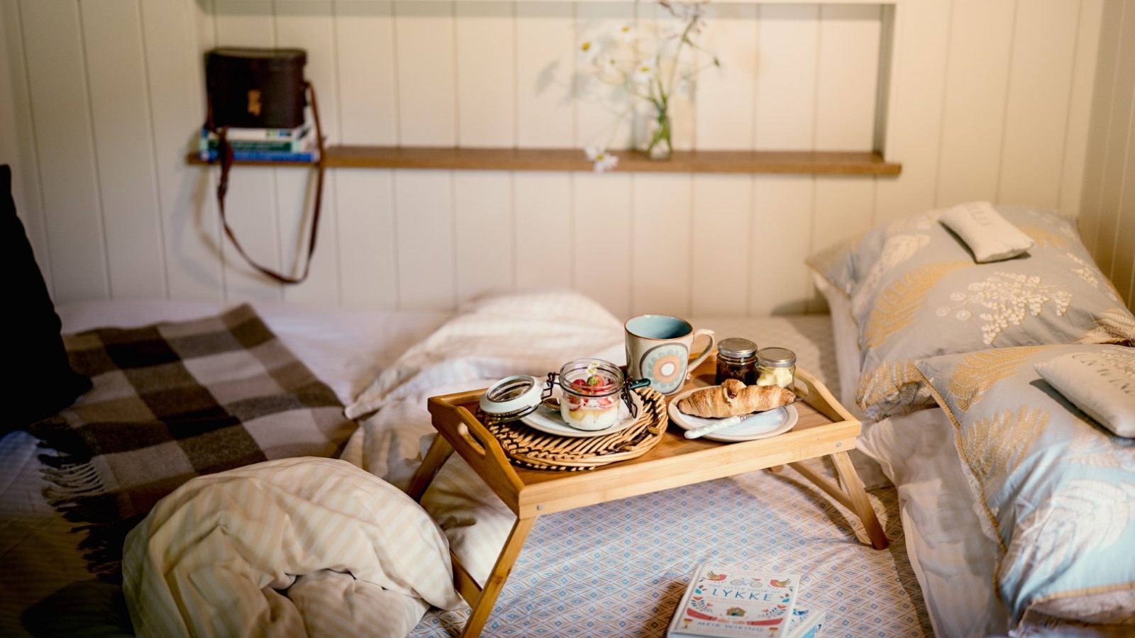 Room with a breakfast tray on a bed, featuring croissants, coffee, and jam. Decor includes books, a blanket, and flower vase.