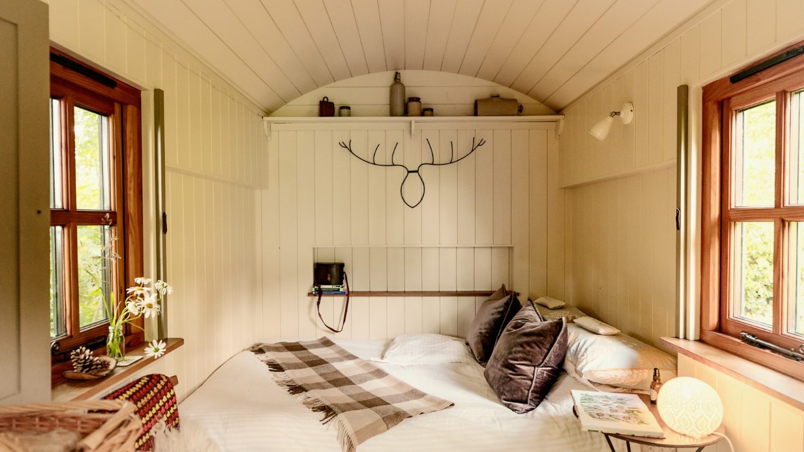 Cozy bedroom with white paneled walls, a double bed with plaid and gray pillows, and a stag antler wire sculpture on the wall.