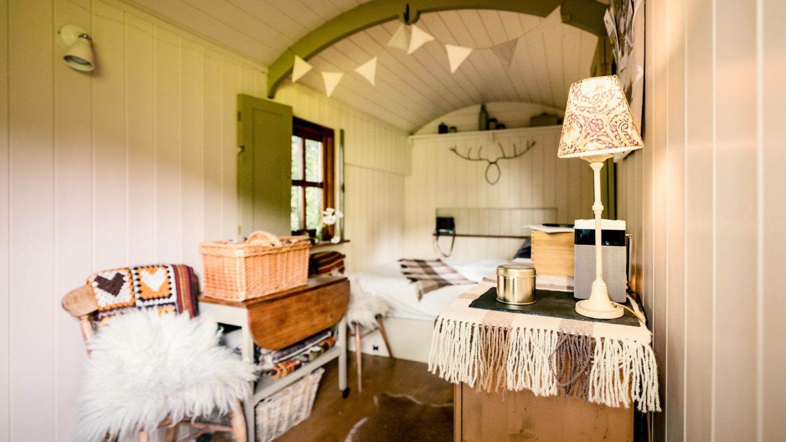 Cozy interior of a small wooden room with a bed, decorated with a basket, a lamp, and wall-mounted antlers.