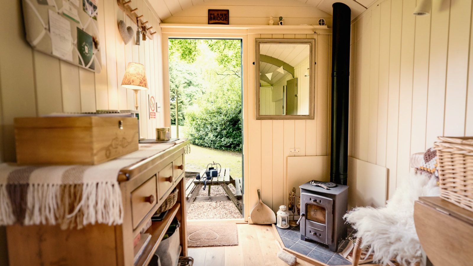 Cozy interior of a small wooden cabin with a wood stove, a chair, and a door open to a lush green garden outside.