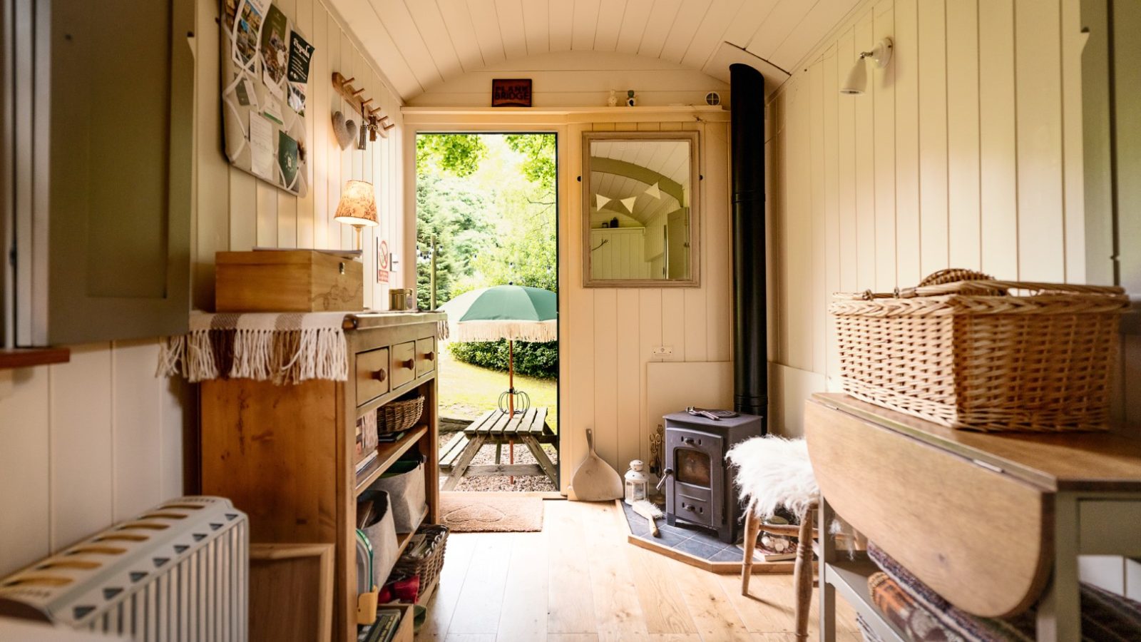 Cozy, rustic interior of a small cabin with a wood stove, shelves, and natural light from an open door leading to a garden.