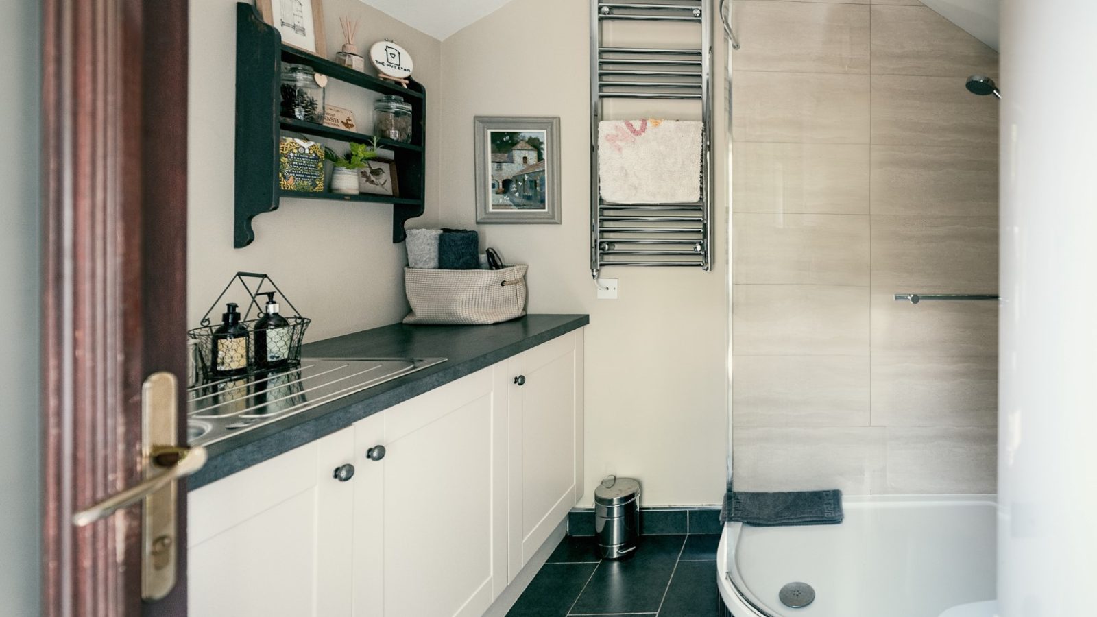 A modern bathroom features a glass shower, countertop with sink, shelves with decor, towel warmer, and a small waste bin.