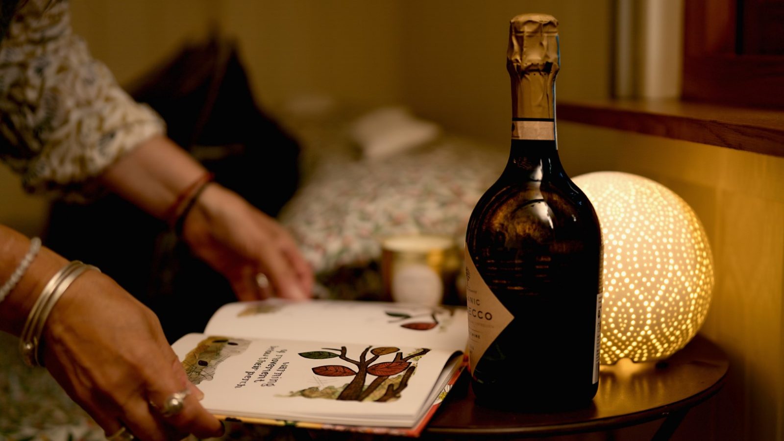 A person reading a book at a bedside table with a bottle of spumante and a glowing lamp.