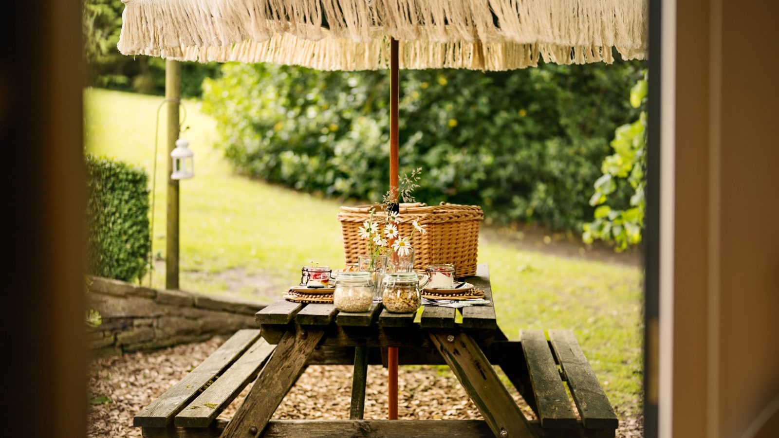 A wooden picnic table with a fringed green umbrella, set with a picnic basket, glass jars, and flowers in a grassy backyard.