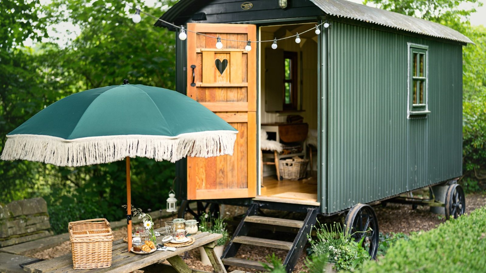 The Hut Eyam is a cosy shepherds hut on wheels with a wooden door, picnic table, umbrella, and string lights in a lush garden setting.