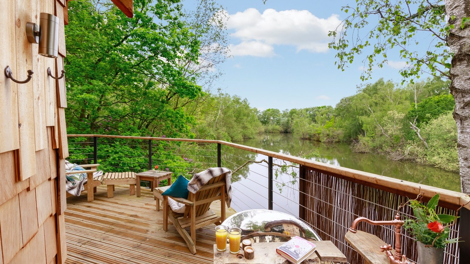 Treetop Hideaway: A wooden deck overlooking a calm river, amidst lush trees, with outdoor seating, drinks, and books on the table.