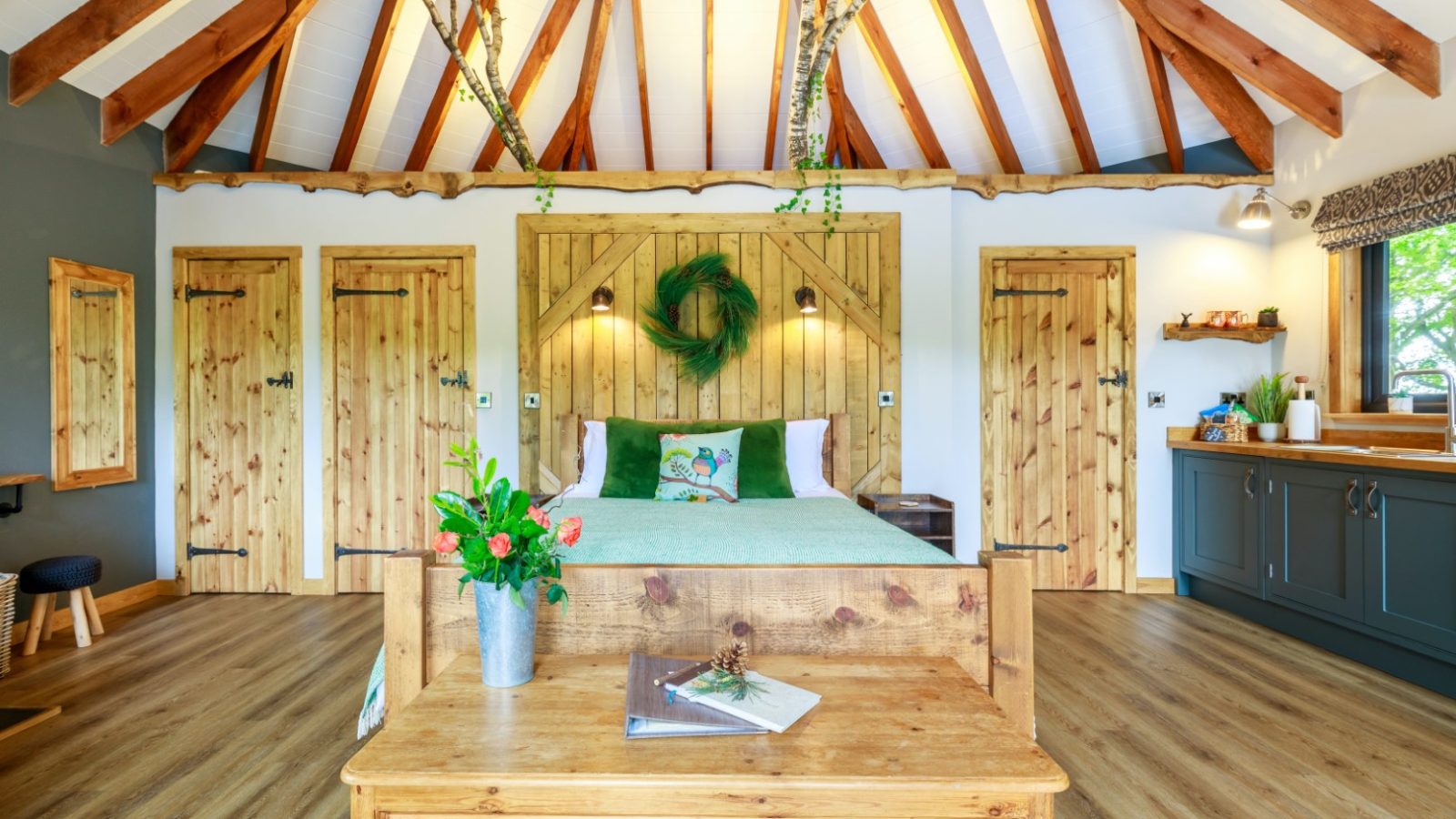 Treetop Hideaway: A rustic bedroom with wooden beams, a wreath, cozy bed, wooden furniture, and a kitchenette on the right.