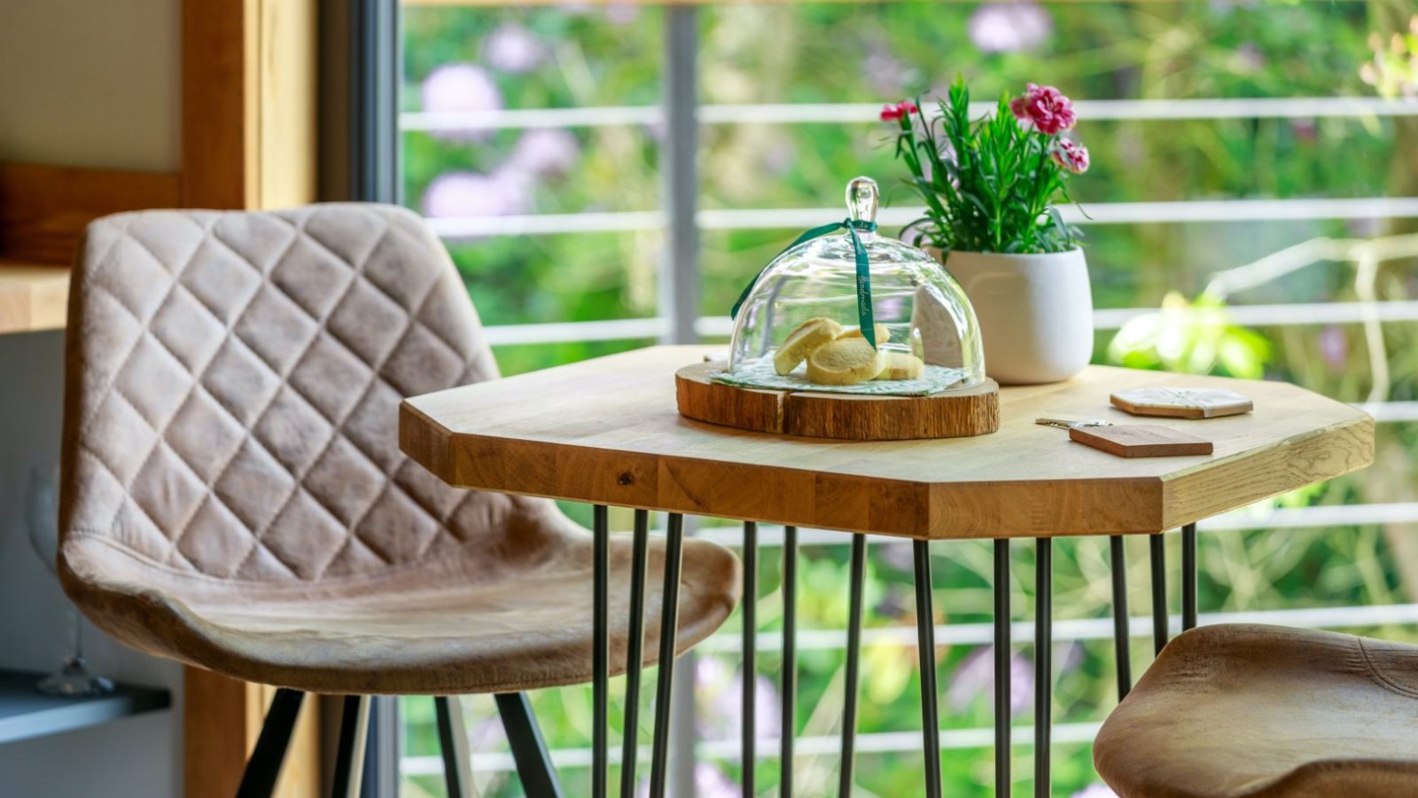 Wooden table by a window in a treetop hideaway, with two chairs, pastries under a glass cloche, plant, and coasters.