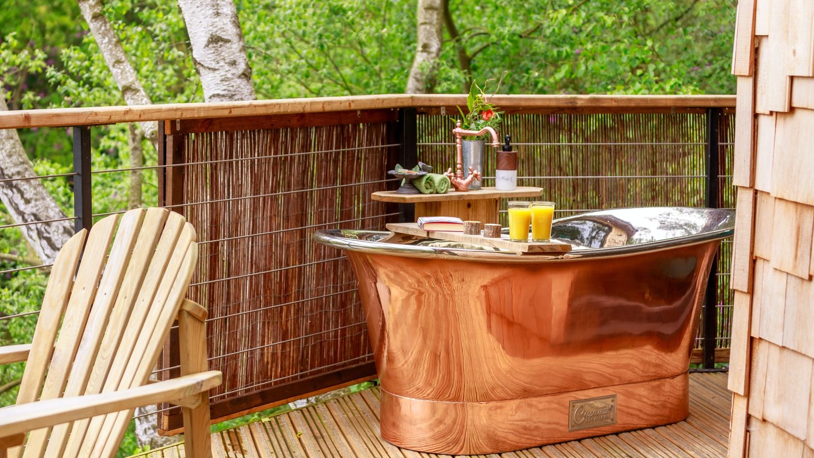 Treetop Hideaway: Copper bathtub on a wooden deck with drinks and flowers, surrounded by greenery and a wooden chair.