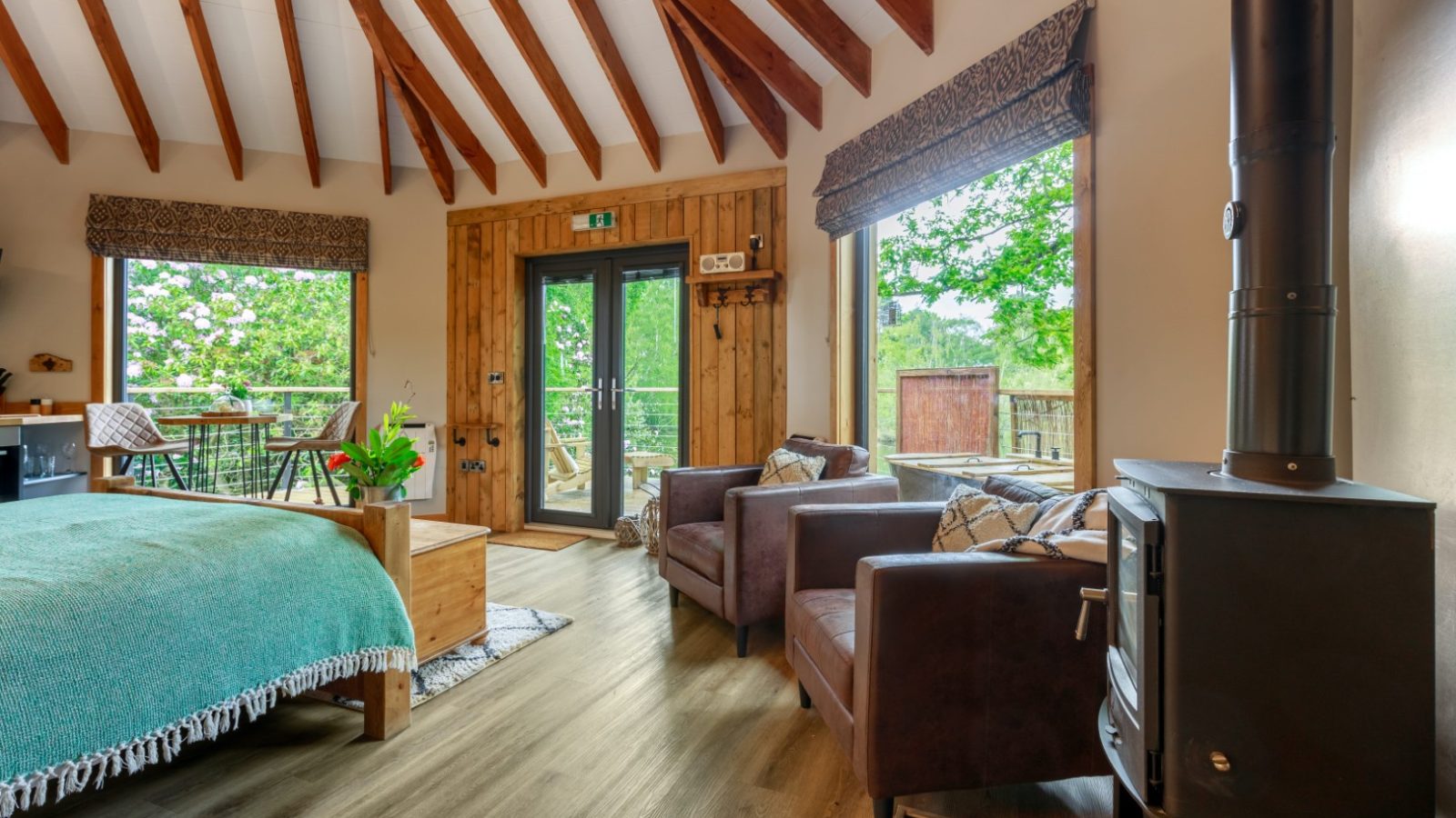 A cozy treetop hideaway with a bed, two armchairs, and a wood-burning stove. Large windows reveal trees outside.