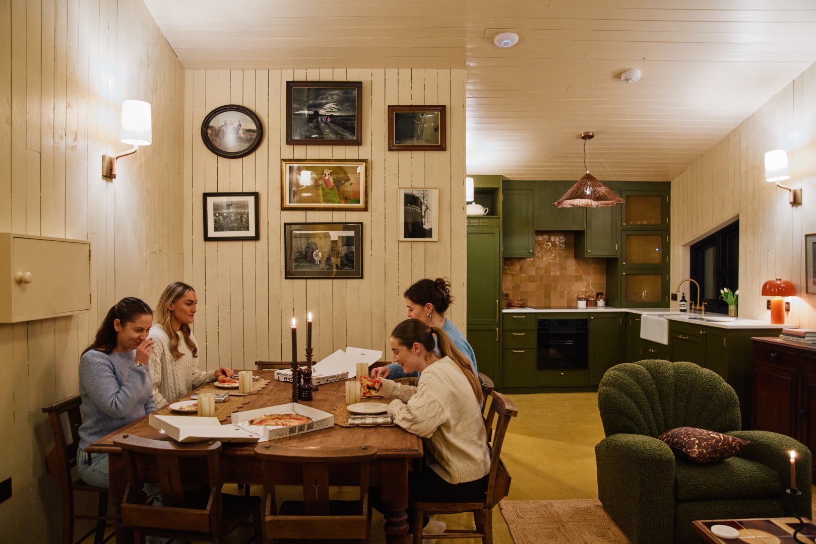 Four friends enjoying pizza at a wooden table, savoring a Girls Weekend Break in a cozy room with framed pictures and a green kitchen.