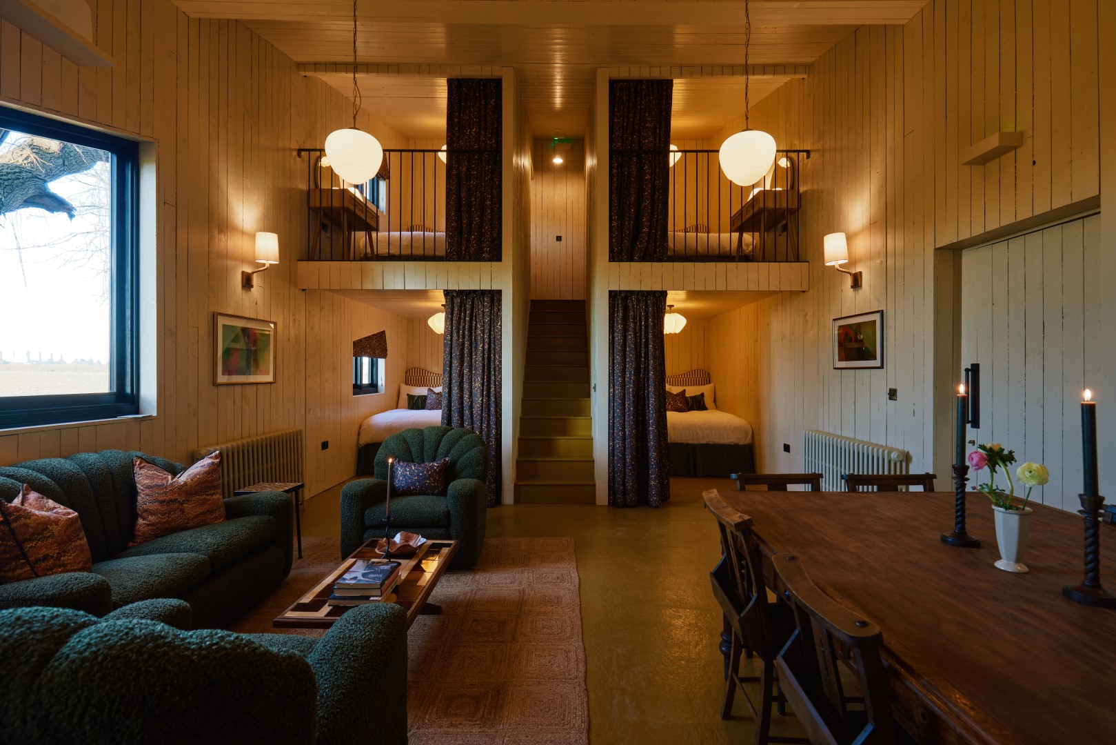 Cozy cabin interior for a girls' weekend break with green sofas, a wooden dining table, and loft beds separated by curtains.