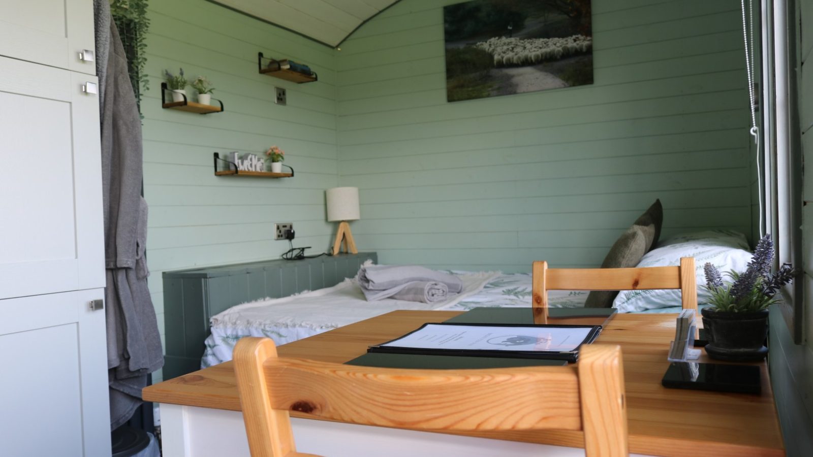 Cozy room with a small wooden dining table, a bed, wall-mounted shelves, and a countryside painting on Breaks Fold Farm green walls.