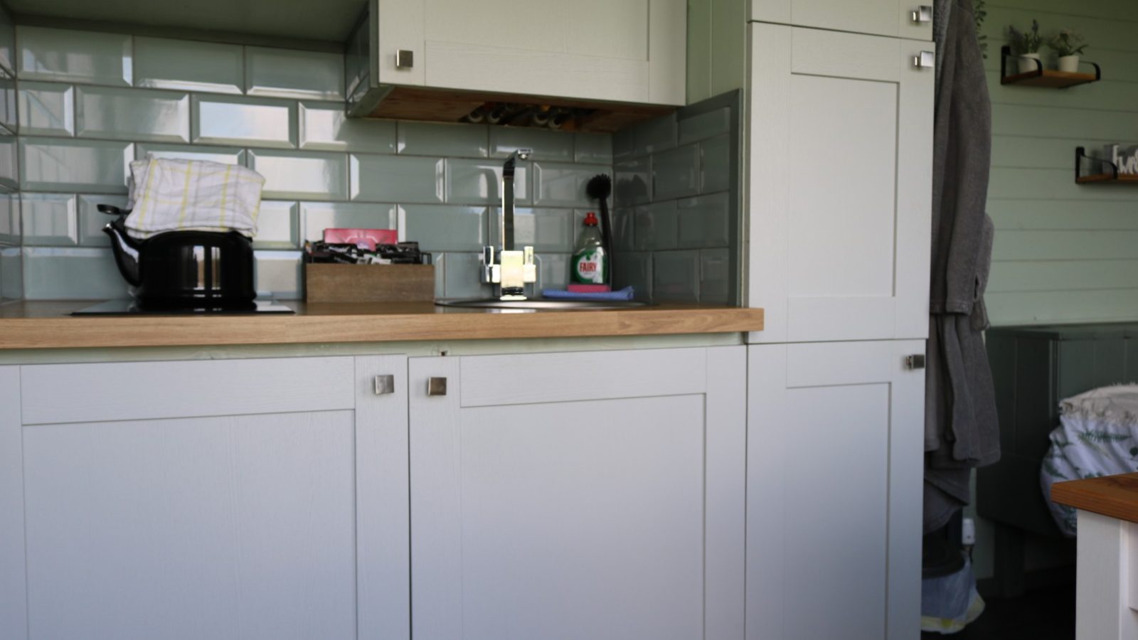 Compact kitchen at Breaks Fold Farm with light gray cabinets, wood countertop, black kettle, and green tile backsplash.