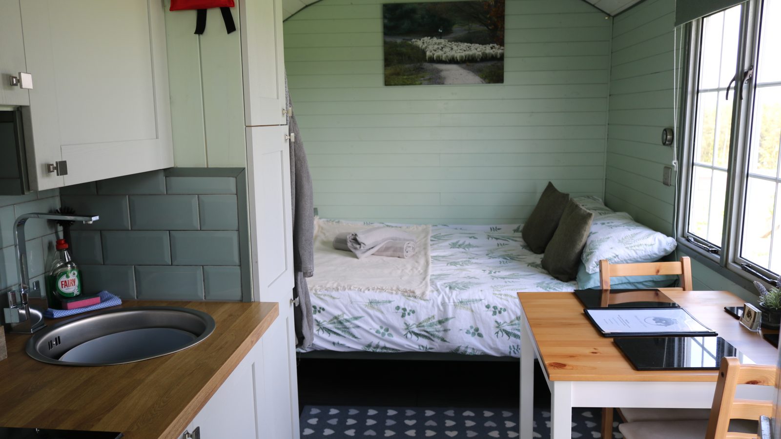 Compact, cozy room with bed, kitchenette, and fold-out dining table. Light green walls with window and landscape artwork.